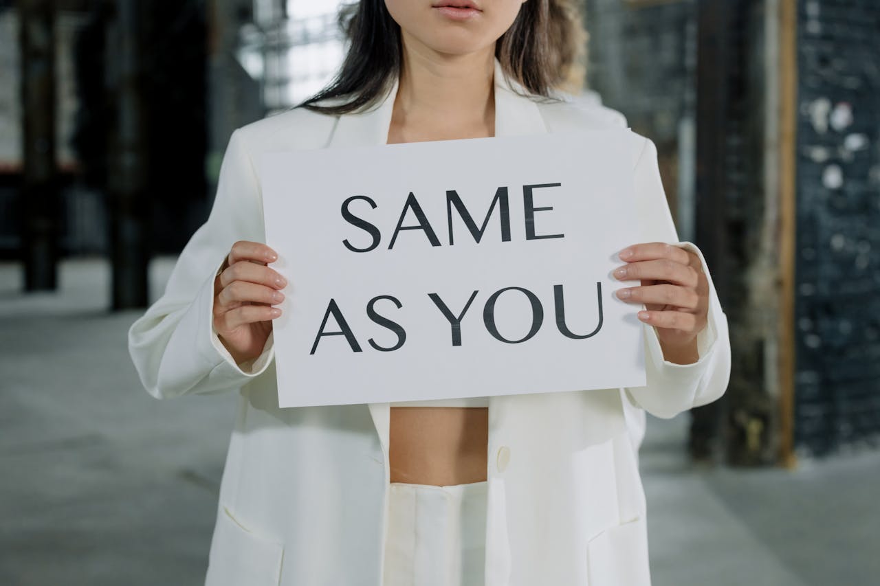 A woman in a white suit holds a sign reading 'Same as You' in an industrial setting.
